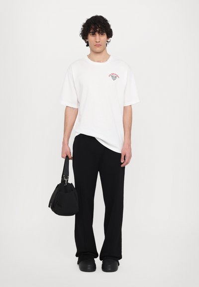 T-shirt blanche à manches courtes avec un imprimé graphique, associée à un pantalon large noir et des chaussures noires. La personne tient un sac noir.