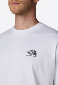 T-shirt bianco in cotone con un logo nero sul lato sinistro del petto che recita "THE NORTH FACE" con la frase "NEVER STOP EXPLORING." Taglio classico.