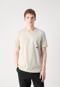 Ung man med ljusbrunt hår som bär en beige kortärmad T-shirt och svarta byxor, stående mot en helt vit bakgrund.