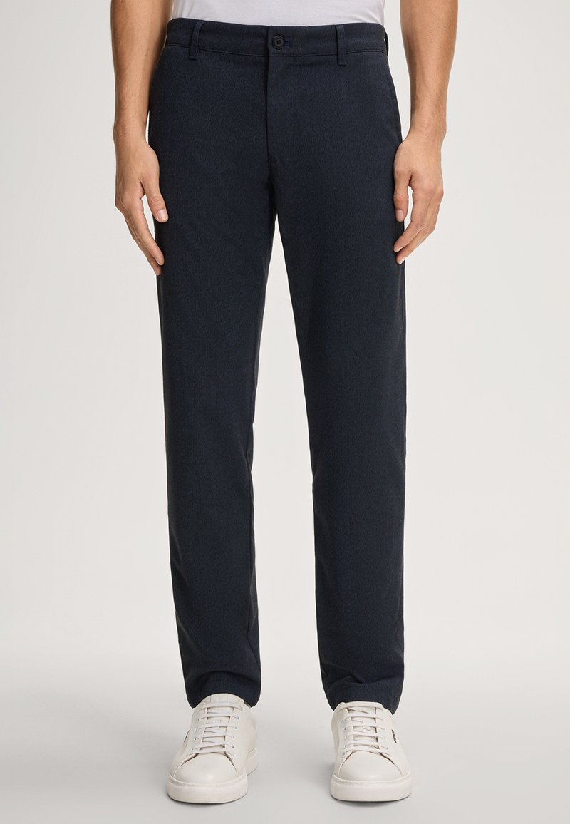 Mężczyzna w ciemnogranatowych spodniach slim-fit i białych sneakersach, stojący na jednolitym jasnym tle.
