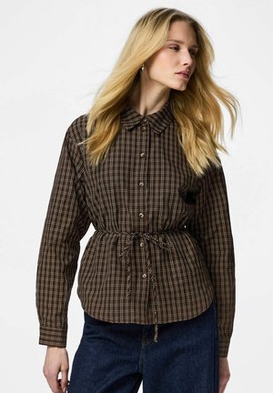 Blond kvinne iført en brun rutete skjorte med knapper og knytebelte i midjen, samt mørkeblå jeans, ser til siden mot en hvit bakgrunn.