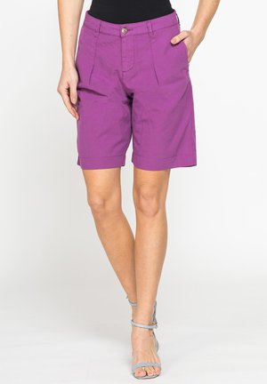 Donna che indossa pantaloncini viola fino al ginocchio, top nero e sandali celesti con cintura alla caviglia, in piedi con una mano in tasca.