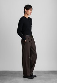 Sweater preto canelado com decote em V, calças castanhas de cintura alta com pregas, cinto preto com fivela dourada, sapatos pretos, de pé sobre um chão cinza.