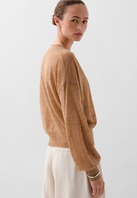Femme avec les cheveux en chignon portant un pull ample marron et un pantalon blanc, regardant par-dessus son épaule sur un fond gris uni.