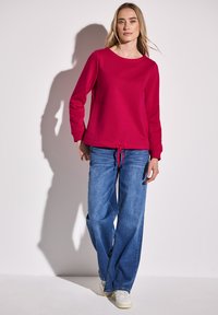 Červená mikina s kulatým výstřihem a dlouhými rukávy, v kombinaci s volnými modrými džíny a bílými teniskami. Měkká látka, volný střih.