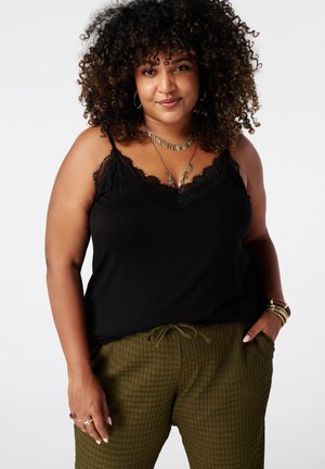 Krullend haar vrouw die een zwart topje met kanten rand draagt, olijfgroene textuurbroek en gelaagde gouden kettingen, staand met handen deels in de zakken.