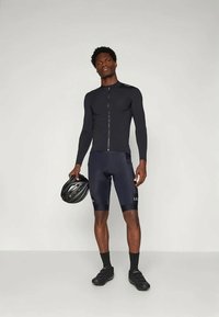 Svart sykkeltopp med lange ermer og glidelås foran, sammen med mørke sykkelshorts. Holder en svart sykkelhjelm.