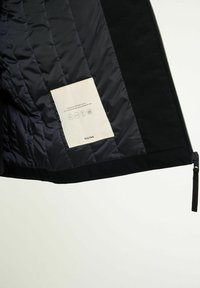 Svart vadderad jackas insida som visar en vit tygmärkning med skötselikoner, text och en dragkedjetab på en ljus enfärgad bakgrund.