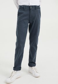 Tmavě modré bavlněné kalhoty se rovnými nohavicemi, ohrnutými manžetami a zapínáním na jeden knoflík vpředu, doplněné bílými teniskami.