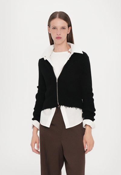 Zwarte geribde cropped zip-up cardigan met een witte gekraagde shirt eronder, met een rafelige zoom, gedragen met bruine tailored broeken.