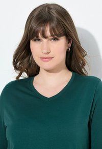 Tmavě zelený top s výstřihem do V vyrobený z hladkého materiálu. Design zahrnuje dlouhé rukávy a přiléhavý střih. Jednoduchý a minimalistický styl.