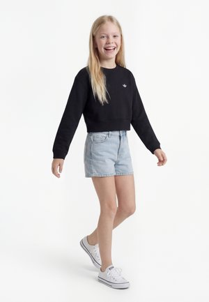 Fille souriante marchant, portant un t-shirt noir à manches longues, un short en denim clair et des baskets blanches sur un fond blanc uni.