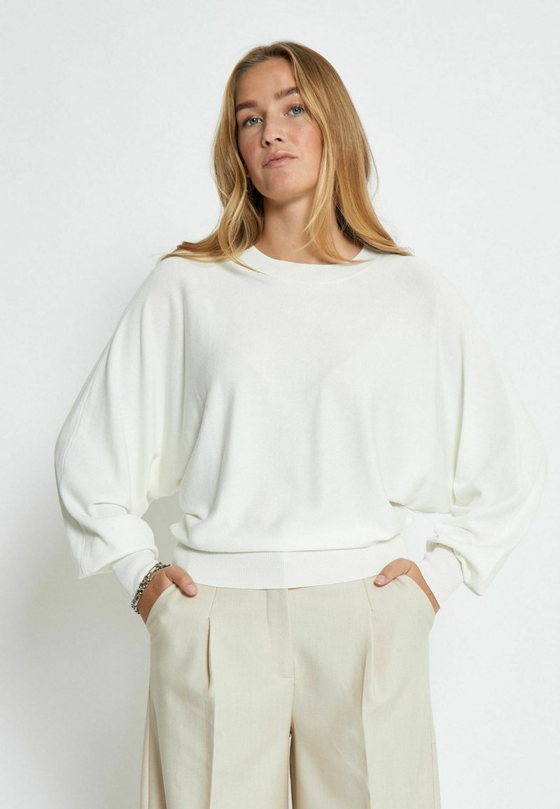 Maglione bianco oversize con scollo a girocollo, spalle scese e polsini a costine, abbinato a pantaloni leggeri beige. Tessuto morbido, vestibilità casual.