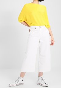 Gelbes geripptes Oversized-Oberteil, weiße weit geschnittene cropped Jeans und weiße High-Top-Sneaker mit roten und blauen Akzenten. Einfaches, lässiges Outfit.
