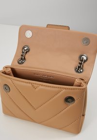 Otevřená béžová prošívaná kožená kabelka se stříbrným řetízkovým popruhem a magnetickým uzávěrem, uvnitř viditelný název značky.