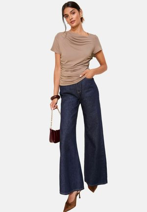 Una modella posa indossando un top beige aderente e jeans larghi scuri, tenendo in mano una piccola borsa bordeaux con tracolla a catena. Tacchi marroni completano l'outfit.