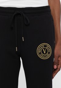 Svarte joggebukser laget av bomullsblanding, med et brodert gullmerke på venstre lår og snormidje med metallender.