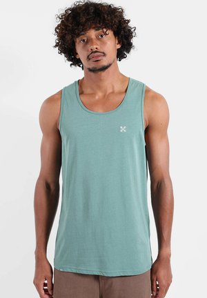 Homme aux cheveux bouclés portant un débardeur turquoise et un pantalon marron, debout devant un fond clair uni.