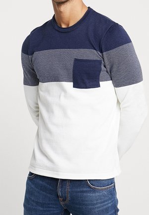 Mann trägt einen Pullover mit rundem Halsausschnitt und langen Ärmeln, mit marineblauen, gepunkteten Streifen, weißen Farbblöcken und einer marineblauen Brusttasche, kombiniert mit blauen Jeans.