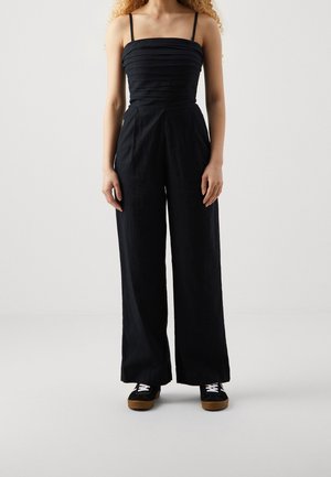 Schwarzer Jumpsuit mit dünnen Trägern, strukturierter, enganliegender Oberteil, weit geschnittene Hose, aus leichtem Stoff, kombiniert mit schwarzen Sneakers.