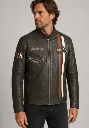 Homme aux cheveux bruns portant une veste de course en cuir noir zippée avec des bandes orange et blanches et des patchs de course, regardant sur le côté.