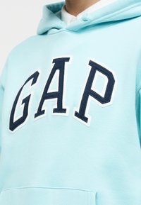 Κοντινό πλάνο ενός ατόμου που φοράει ανοιχτό μπλε φούτερ με κουκούλα, με μεγάλα κεντημένα γράμματα «GAP» σε σκούρο μπλε και λευκό στο μπροστινό μέρος.