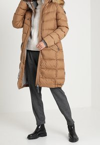 Abrigo hinchado marrón con capucha forrada de piel, bolsillos con cremallera, textura acolchada y un diseño largo. Combinado con pantalones grises y botas negras brillantes.