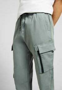 Man som bär ljusgröna cargobyxor med dragsko i midjan och dragkedjefickor på sidan, tillsammans med en vit skjorta.