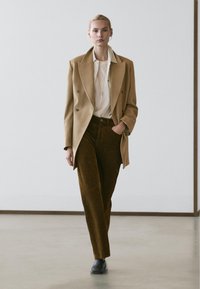 Kvinde går indendørs iført en beige dobbeltradet blazer, stribet skjorte, hvid top, brune bukser og sorte sko, med den ene hånd i lommen.