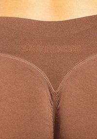 Braune Leggings mit geripptem Bund und eingeprägtem Markennamen. Weiche Textur und figurbetontes Design. Nahtdetails betonen die Form.