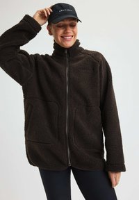 Nainen hymyilee päällään tummanruskea vetoketjupusero ja musta lippalakki, yksi käsi säätelee lippalakkia vaaleaa taustaa vasten.