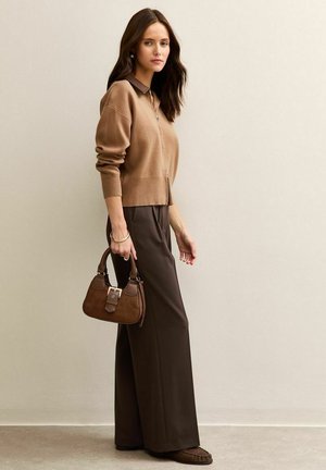 Cardigan marrón de punto con cremallera, combinado con pantalones marrones de pierna ancha. Sosteniendo un pequeño bolso marrón con cierre y detalles texturizados.