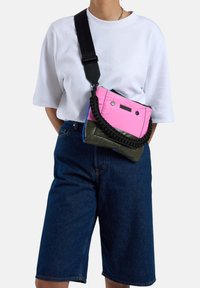 Kleurvolle nylon schoudertas met roze en olijfgroene secties, gevlochten handvat en een ritssluiting met twee accenten knopen.