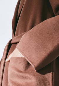 Cappotto in morbido tessuto marrone con colletto a scialle, maniche ampie e tasca laterale. La texture sembra liscia e soffice.