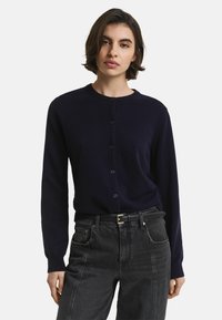 Marinblå cardigan med rund hals, långa ärmar och knäppning fram. Tillverkad av ett mjukt material, bärs över högmidjade svarta jeans.