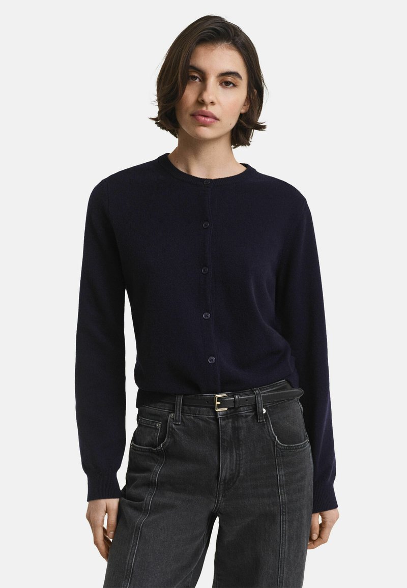 Marinblå cardigan med rund hals, långa ärmar och knäppning fram. Tillverkad av ett mjukt material, bärs över högmidjade svarta jeans.