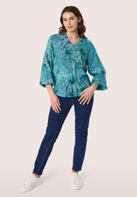 Turquoise bloes met bloemmotief, V-hals, en klokmouwen; gecombineerd met donkerblauwe skinny jeans en witte sneakers. Soepele textuur en losse pasvorm.