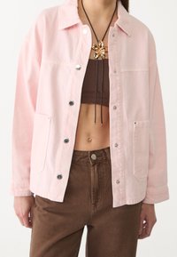 Chaqueta de mezclilla rosa con botones plateados, dos bolsillos frontales y un corte relajado. Llevada sobre un top corto marrón y combinada con pantalones marrones.