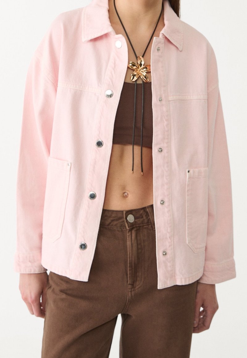 Chaqueta de mezclilla rosa con botones plateados, dos bolsillos frontales y un corte relajado. Llevada sobre un top corto marrón y combinada con pantalones marrones.