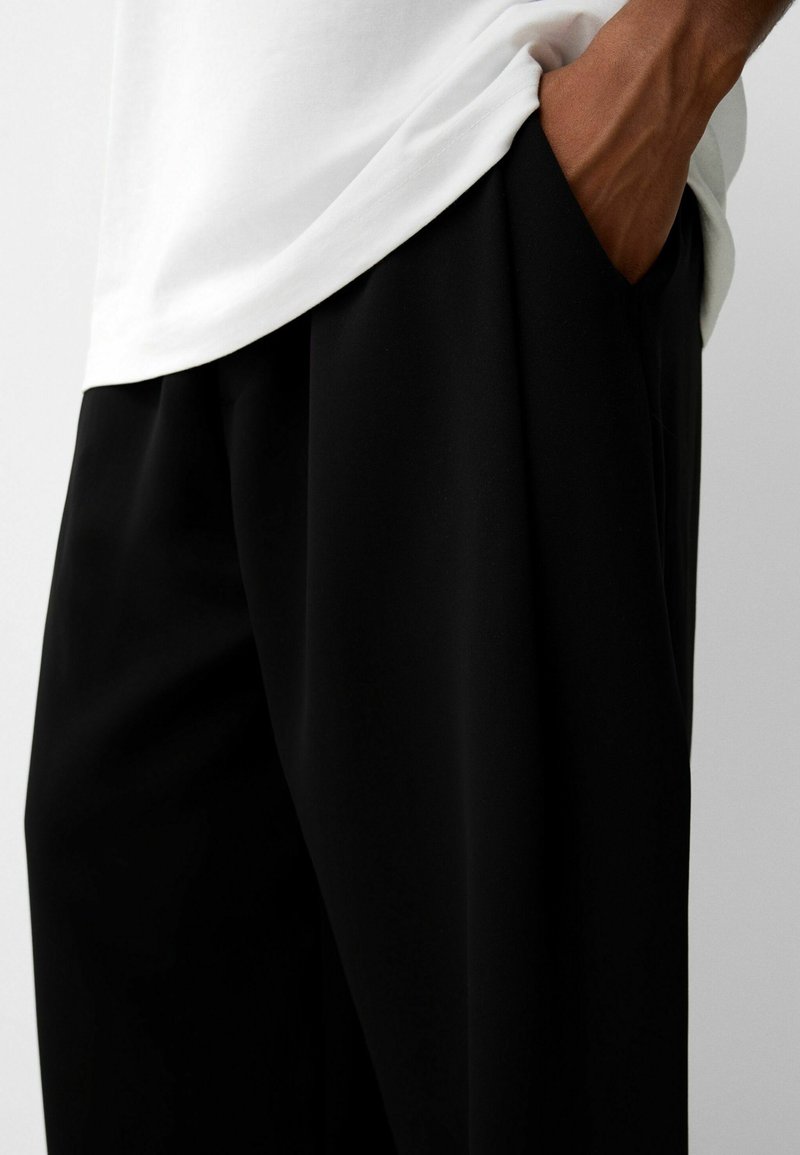 Czarne, luźne spodnie wykonane z lekkiego materiału, z szerokimi nogawkami i boczną kieszonką, w połączeniu z białą koszulą.