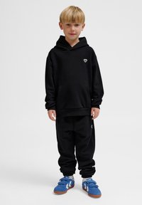 Svart huvtröja med framficka och logotyp; svarta sweatpants; blå sneakers med kardborreband och vita detaljer.