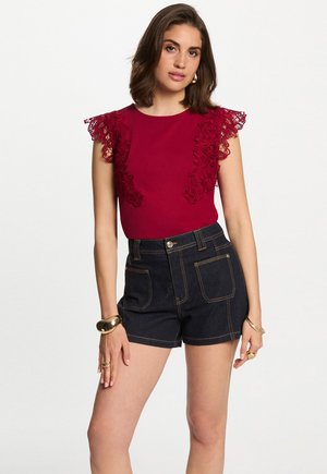 Top bordeaux con dettagli in pizzo sulle maniche, abbinato a pantaloni corti di jeans scuri a vita alta con cuciture a contrasto. Tessuto dalla consistenza liscia.