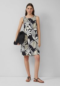 Zwarte jurk met crèmekleurige bladenprint, mouwloos ontwerp en een relaxte pasvorm, gecombineerd met een gestructureerde zwarte tas en platte zwarte sandalen.