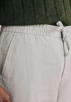 Henkilö, jolla on päällään vaaleanbeiget narulla kiristettävät pellavahousut ja tummanvihreä ribbineulottu neulepusero, käsi lepäämässä taskun lähellä.