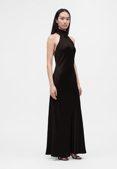 Femme aux longs cheveux foncés portant une robe noire sans manches, à col montant et longue jusqu'au sol, ainsi que des talons noirs à bouts ouverts, debout devant un fond blanc.