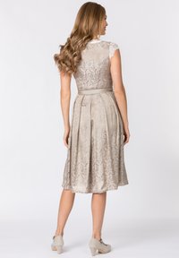 Ein knielanges, hellbeiges Kleid mit strukturierter Spitze, plissiertem Rock und kurzen Ärmeln, kombiniert mit grauen Absatzschuhen.