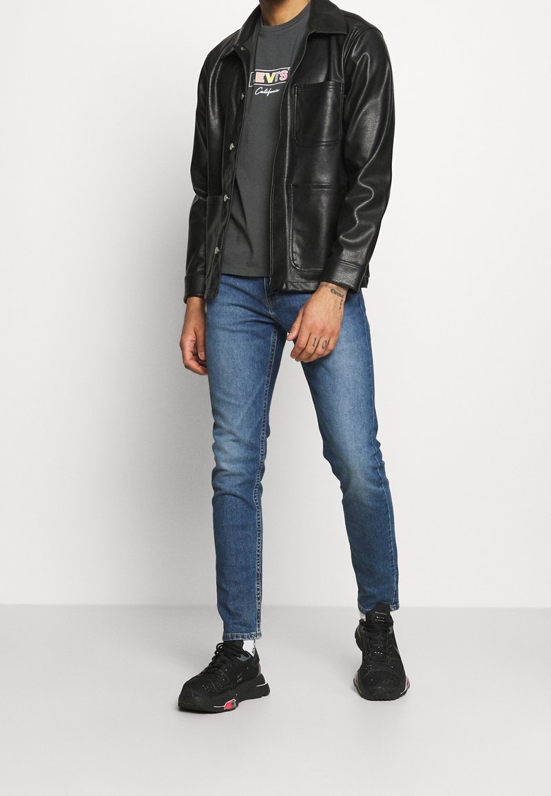 Homme portant une veste en cuir noire, un t-shirt Levi’s gris foncé, un jean bleu et des baskets noires sur un fond blanc uni.