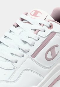 Białe skórzane sneakersy z różowymi akcentami, okrągły czubek, płaskie sznurowadła, wyściełany język oraz detal z logo po boku. Widoczne teksturowane szwy.