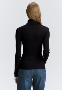 Pull en maille côtelée noir à col roulé avec des manches longues, présentant une coupe ajustée et un tissu texturé. Jeans en denim visibles dans la partie inférieure.