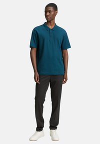 Zöldeskék póló klasszikus gallérral és három gombbal, sötétszürke nadrággal és fehér sportcipővel párosítva, lazább fazonban.